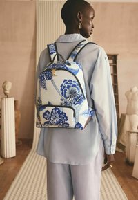 Mochila azul y blanca con un patrón floral, con correas ajustables y un bolsillo delantero. Hecha de tela, presenta una textura suave y herrajes dorados.