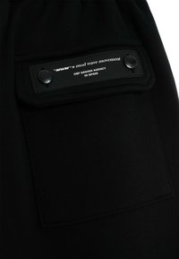 Joggers de tela negra con un bolsillo lateral, un parche de cuero rectangular, logo en relieve y detalle de botón a presión.