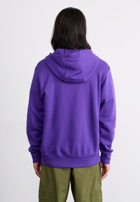 Corta-vento roxo com capuz ajustável, punhos e barra canelados. O tecido parece suave e semelhante a fleece, com um corte mais descontraído.