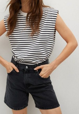 Vrouw draagt een mouwloos wit topje met zwarte horizontale strepen en hooggetailleerde zwarte denim shorts met handen in haar zakken.