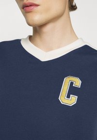 Tmavě modré tričko s výstřihem do V, krémovým lemováním a vyšívaným zlatým písmenem "C" na hrudi. Bavlněná látka s hladkým povrchem.