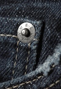 Fecho de mola metálico com as letras gravadas "WEEK DAY" em tecido de ganga azul escuro, com costura branca e textura visível.