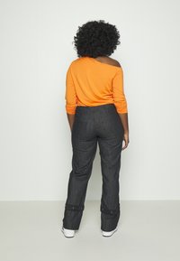Orange långärmad topp med off-shoulder-design och gathered ärmar, parat med högmidjade mörk denimbyxor med tryckknappar.