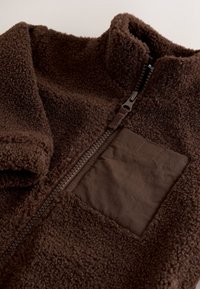 Veste en polaire marron avec un col haut, dotée d'une fermeture éclair à l'avant et d'une poche à rabat en tissu lisse de couleur marron plus foncée. Texture douce.
