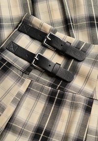 Tissu à carreaux beige avec des lignes noires et grises, doté de deux sangles en cuir noir et de boucles argentées pour l'ajustement.