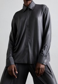 Person som bär en löst sittande, mörkgrå satin skjorta med knappar och breda manschetter, samt matchande vida byxor mot en grå bakgrund.