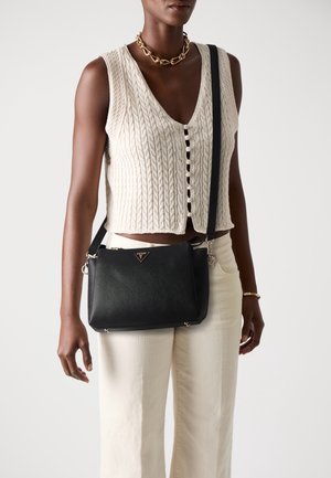 Borsa a tracolla nera con finitura testurizzata, hardware in tono oro e accento del logo. Indossata con un top in maglia color crema e pantaloni chiari.