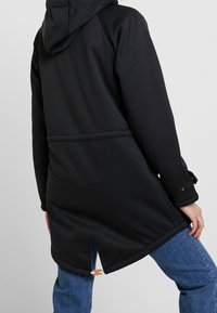 Veste noire à capuche avec un design épuré, tissu texturé et détail de ventilation dans le dos, assortie à un jean en denim.