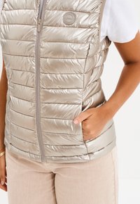 Metaliczna beżowa kamizelka puchowa z poziomymi przeszyciami, zapięciem na zamek i bocznymi kieszeniami. Posiada okrągłą naszywkę z logo na klatce piersiowej.