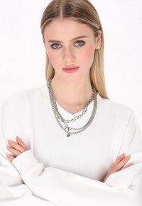 Kerangkai rantai perak dengan ketebalan yang bervariasi. Model mengenakan sweatshirt putih, dengan tangan disilangkan, menonjolkan tekstur dan kilau perhiasan.