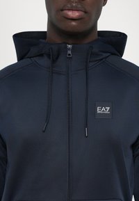 Hoodie zippé bleu marine avec une finition texturée, dotée d'une capuche avec cordon de serrage et d'un patch logo EA7 sur la poitrine. Tissu lisse et design moderne.
