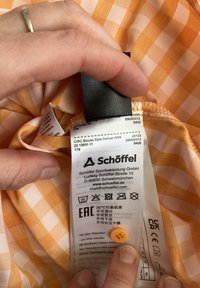 Orange-weißer kariert Stoff mit einem Pflegeetikett, das schwarzen Text enthält. Das Etikett umfasst Waschhinweise und einen abnehmbaren Knopf.