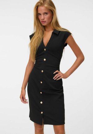 Femme blonde portant une robe noire sans manches moulante avec des boutons dorés sur le devant, debout sur un fond blanc.