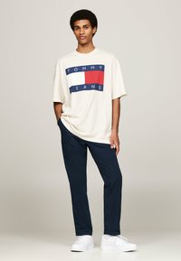 Camiseta oversized color crema con un diseño gráfico en azul, rojo y blanco en la parte frontal, combinada con pantalones azul marino y zapatillas blancas.