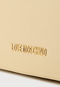 Μπεζ υφασμάτινη επιφάνεια δέρματος με χρυσό χρώμα λογότυπου "LOVE MOSCHINO" σε πρώτο πλάνο, με λεπτομέρειες ραφής.