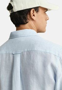 Hombre vistiendo una camisa de lino azul claro y una gorra blanca de béisbol, de perfil con atención en el detalle de la espalda y el cuello de la camisa.