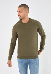 Sudadera de cuello redondo en color verde oliva, confeccionada en una tela suave. Presenta puños y dobladillo con canalé, además de una textura lisa. Combinada con unos jeans de color azul claro.
