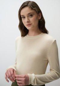 Maglione lavorato a maglia di colore crema, con maniche lunghe, colletto e polsini a coste. Materiale morbido e leggero con sottili dettagli di texture.