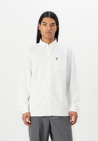 White button-up shirt made of cotton, featuring a chest pocket and a yellow embroidered logo. Long sleeves with button cuffs.