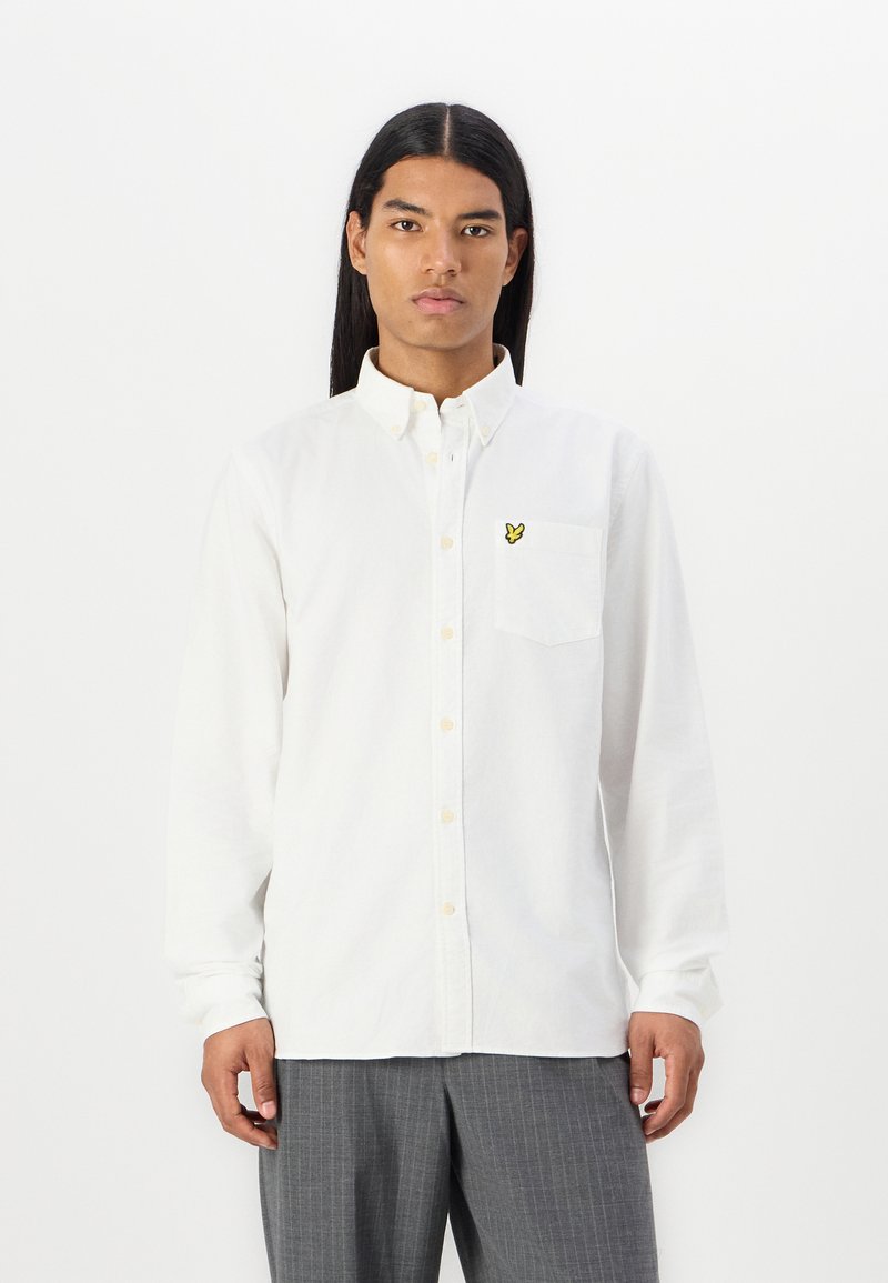 White button-up shirt made of cotton, featuring a chest pocket and a yellow embroidered logo. Long sleeves with button cuffs.