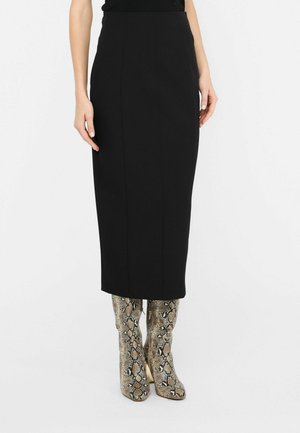 Mujer que lleva una falda lápiz midi negra combinada con botas altas hasta la rodilla con estampado de piel de serpiente, de pie frente a un fondo blanco.