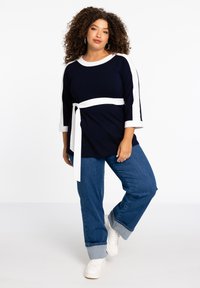 Marineblauer Oberteil mit weißen Ärmeln und einem Bindegürtel, kombiniert mit blauen Jeans und weißen Sneakers, bietet einen lässigen Schnitt und eine weiche Stoffstruktur.