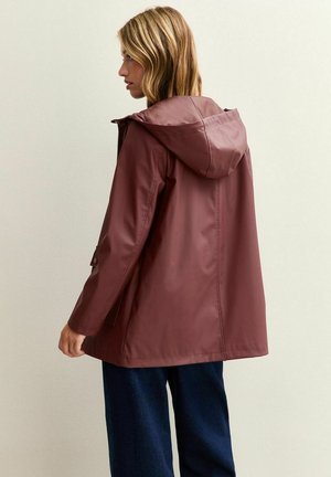 Femme aux cheveux blonds portant un manteau de pluie amples de couleur bordeaux avec capuche et un pantalon bleu foncé, tournée vers la gauche sur un fond clair et uni.