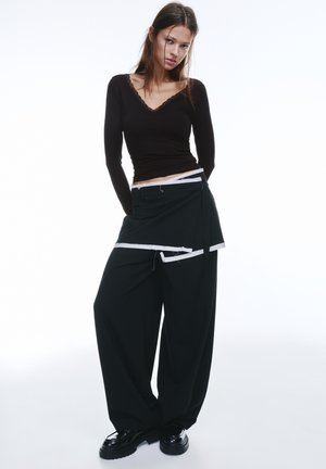 Zwarte wijde broek met een gelaagd, asymmetrisch ontwerp en witte accenten. Fitted top met lange mouwen in donkerbruin met kanten afwerking.