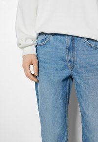 Jeans de mezclilla azul claro con un corte relajado, que presentan bolsillos frontales y costuras visibles. Llevados con una camisa blanca de manga larga.