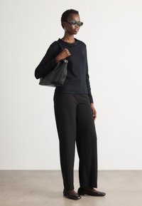 Zwarte lange mouwen top, zwarte brede broek, zwarte platte schoenen en een zwarte handtas met gouden hardware. Model draagt donkere zonnebril.