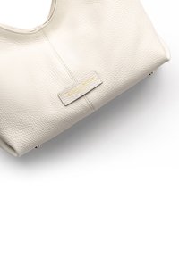 Cremfarbene Lederhandtasche mit strukturiertem Finish, ausgestattet mit einer genähten Naht und einem kleinen rechteckigen Anhänger mit goldener Schrift. Abgerundete Form mit flachem Boden.