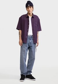 Πουκάμισο με κοντά μανίκια και καρό σχέδιο σε σκούρο μωβ και κόκκινο, φορεμένο πάνω από λευκή αμάνικη μπλούζα. Χαλαρά, ανοιχτόγαλανά τζιν παντελόνια και μαύρα αθλητικά παπούτσια ολοκληρώνουν το σύνολο.