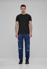 Svart kortermet t-skjorte, blå slitte jeans med lyse flekker, og svarte ankelstøvler. Modellen står oppreist mot en grå bakgrunn.