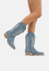 Botas de vaquero de mezclilla azul claro con bordado floral, caña de altura media, punta afilada y tacón de bloque de madera. Cierre de cremallera en la parte posterior.