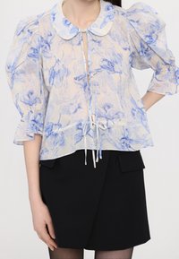 Lekka, przezroczysta bluzka z niebieskim kwiatowym wzorem, z kołnierzykiem typu Peter Pan i bufiastymi rękawami. Zawiązywana z przodu, o luźnym kroju.