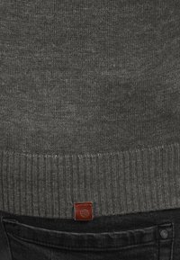 Grå strikket sweater med ribbet afslutning, der har en struktureret overflade og et lille logo i brunt læder. Matchet med sorte jeans.
