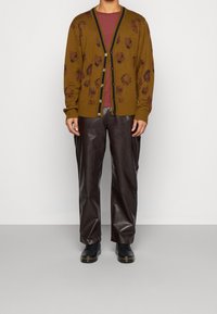 Cárdigan marrón de punto con un patrón gráfico, con ribetes en negro y botones, combinado con pantalones de cuero marrón oscuro y zapatos negros.
