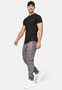 Sort tætsiddende t-shirt, lys grå cargo-bukser med flere lommer og hvide klumpede sneakers. Modellen bærer også en sort kasket.