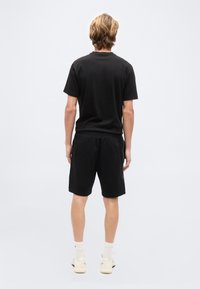 Άνδρας με ξανθά μα λλιά, ντυμένος με μαύρο μπλουζάκι, μαύρο σορτς, λευκές κάλτσες και λευκά αθλητικά παπούτσια, στέκεται με την πλάτη προς τα εμπρός σε απλό φόντο.