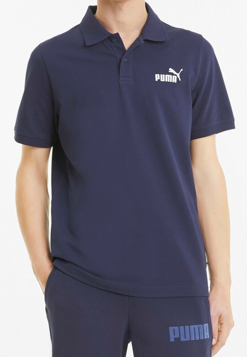 Homme portant un polo bleu marine Puma et un pantalon assorti bleu marine avec le logo Puma sur la poitrine et la cuisse.
