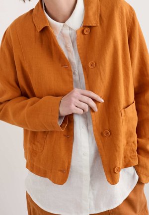 Persona abotonando una chaqueta color naranja quemado sobre una camisa blanca con cuello, mostrando tres botones visibles y bolsillos delanteros.