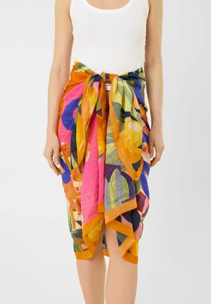 Persoon draagt een veelkleurige bloemige sarong, in de taille gestrikt over een wit tanktop, staand tegen een effen achtergrond.