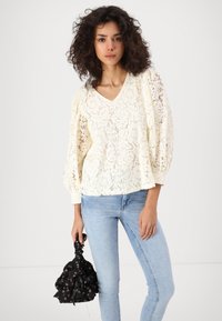 Kvinne med krøllete hår som har på seg en kremfarget bluse med blonder og lys blå jeans, holder en svart veske med blomsterdesign.