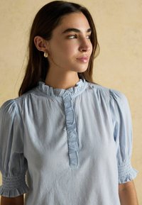 Junge Frau mit langen dunklen Haaren, die eine hellblaue Bluse mit Nadelstreifen und Rüschensaum sowie -Ärmeln trägt und seitlich vor einem beigen Hintergrund schaut.