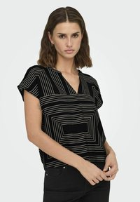 Blusa nera a maniche corte con scollatura a V profonda, caratterizzata da motivi lineari bianchi che creano un design geometrico. Tessuto morbido, vestibilità rilassata.