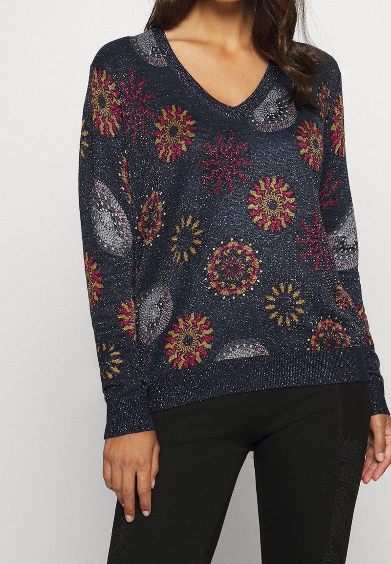 Femme portant un pull à col en V bleu marine scintillant avec des motifs circulaires et en spirale colorés en rouge, or et gris, associé à un pantalon noir.