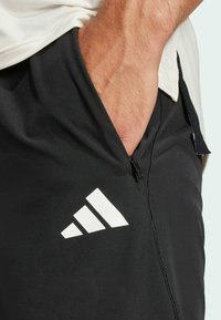 Svarta träningsoveraller med en slät yta, utrustad med en dragkedjeficka och en vit Adidas-logotyp på nedre vänstra sidan.