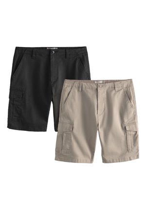 Ung mand står med hænderne i lommerne iført hvid kortærmet skjorte, sorte shorts, beige sweater over skuldrene, beige sokker og hvide sneakers.