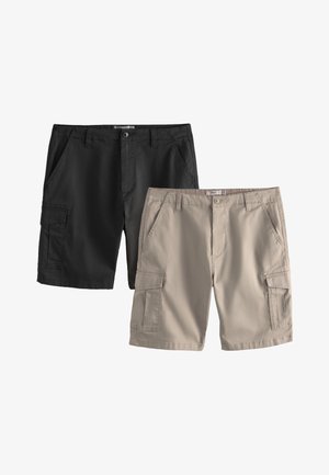 Joven de pie con las manos en los bolsillos, lleva una camisa blanca de manga corta, pantalones cortos negros, un suéter beige sobre los hombros, calcetines beige y zapatillas blancas.
