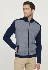 Sweat à fermeture éclair avec un devant en tricot texturé gris clair et des manches et un col bleu marine unis. Comprend des poches latérales et des poignets côtelés.